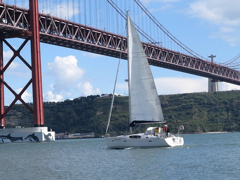 A sailboat passing under the red steel deck of the bridge